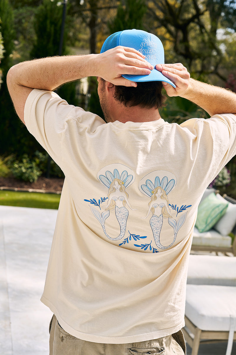 A person models The Siren Collection: SDS Sirens Tee, a light beige unisex T-shirt featuring a blue mermaid design on the back. They hold a blue cap amid greenery and outdoor furniture, enhancing the retro vibe.