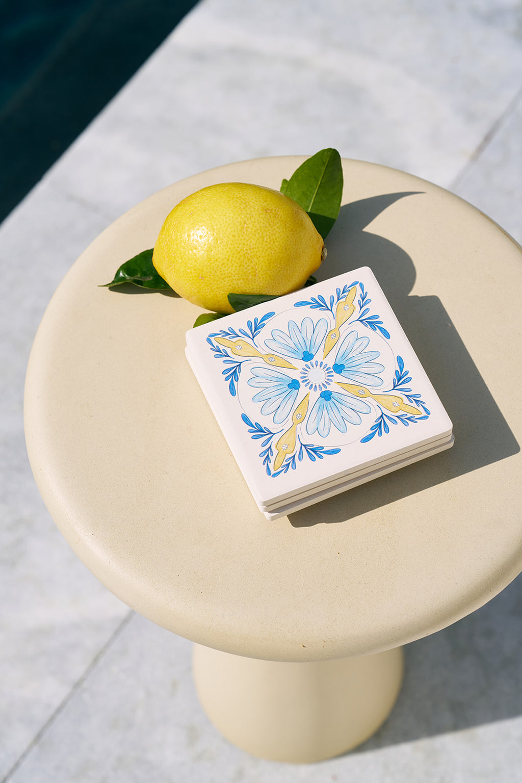 A lemon with leaves sits on a light-colored round table next to Euro Blue Tile Coasters, featuring blue and yellow floral patterns with slip-resistant cork bases. A dark-colored pool is visible in the background.