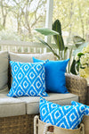 A cozy patio with a light beige sofa, vibrant Blue Ikat Pillow, two blue-patterned pillows, a solid blue pillow, a woven basket holding another blue-patterned pillow, green plants, and white flowers in the background.