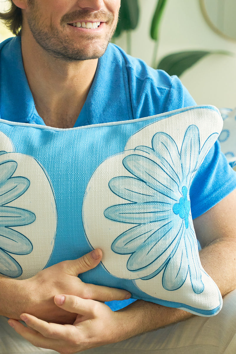 A smiling man in a blue shirt holds the Calico Scallop Lumbar Pillow, featuring an elegant floral design. The softly blurred, green background enhances the relaxed, coastal summer vibe of this sophisticated accent piece.