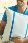 A person in a blue shirt smiles while holding the Line of Euro Blue Tiles Pillow, a white cushion accented with two bright blue vertical stripes and blue piping, evoking the coastal charm of Mediterranean villages.