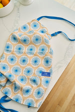 A yellow Calico Scallops Apron with blue flower patterns and an adjustable neck strap lies on a white marble countertop next to a bowl of oranges. Inspired by Sewing Down South patterns.