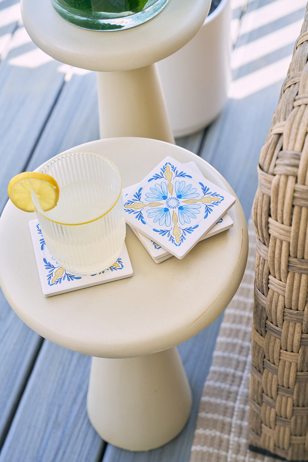 A glass of lemonade with a lemon slice rests on a white table beside the Euro Blue Tile Coaster Set, next to a woven chair and beige rug on a wooden deck.