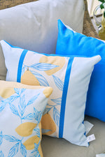 Three decorative pillows on a light gray outdoor seat: the Lemons in a Line Pillow featuring lemons and blue leaves, a solid blue pillow, and one with a partial lemon design. Wicker texture in the background enhances the citrus-inspired decor.