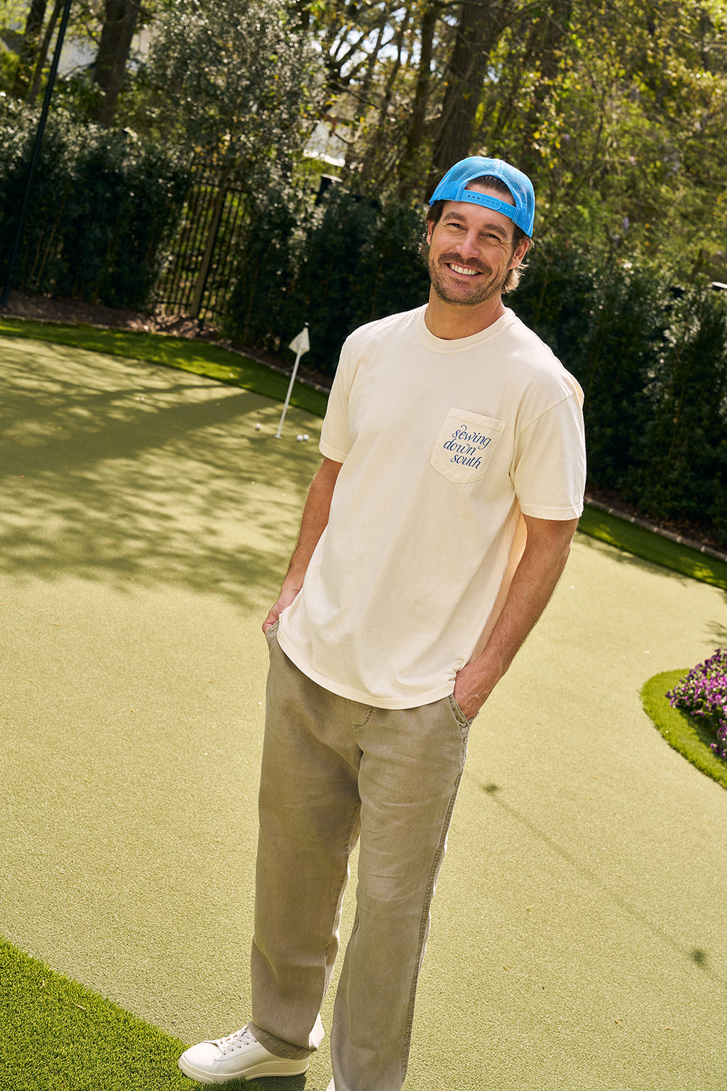 A man in a blue cap, The Siren Collection: SDS Sirens Tee, and khaki pants smiles on a sunny outdoor putting green, surrounded by trees and bushes.