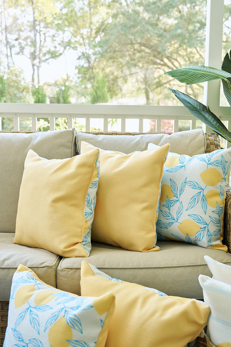A beige outdoor sofa forms a cozy spot with yellow pillows and a Lots of Lemons Pillow adorned with blue leaves. Sunlight pours in, while greenery is visible beyond the white railing in the background.
