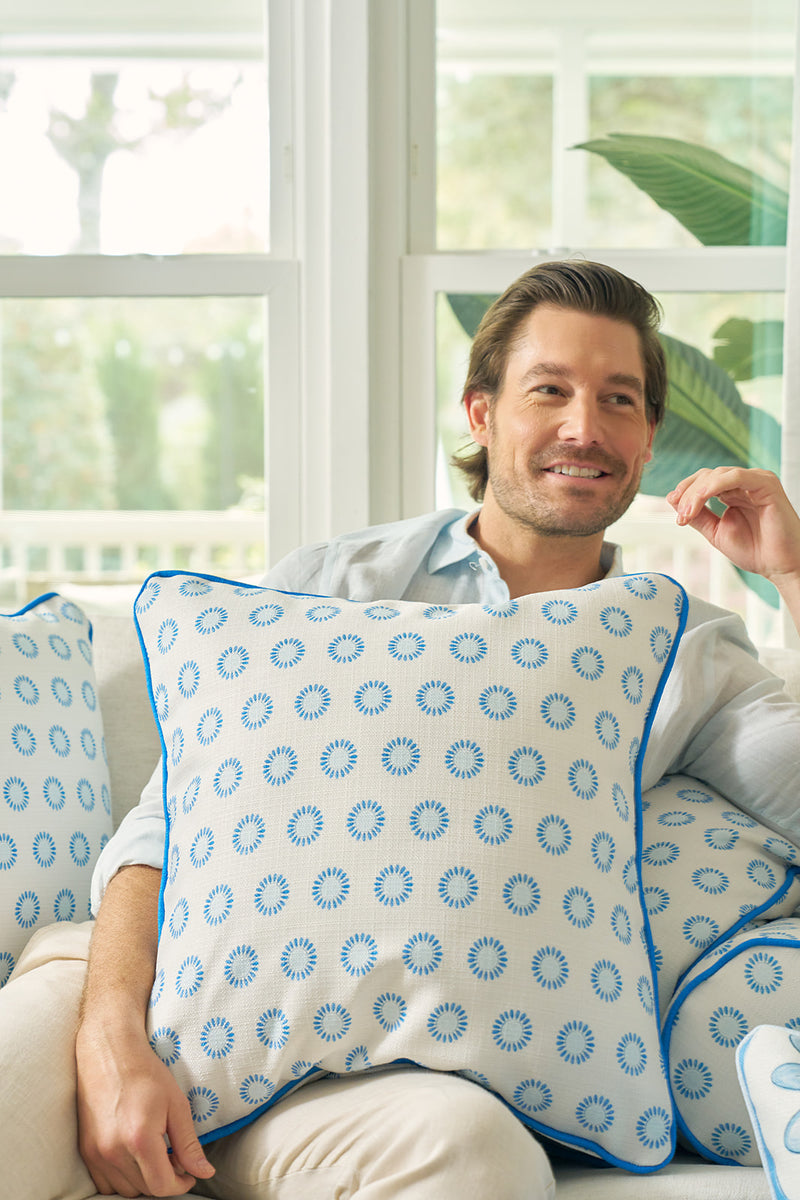 A man with light brown hair in a light blue shirt smiles on a couch, holding a Calico Mini Floral Oversized Pillow with bright blue piping, surrounded by similar pillows. Large windows and plants are visible in the background.