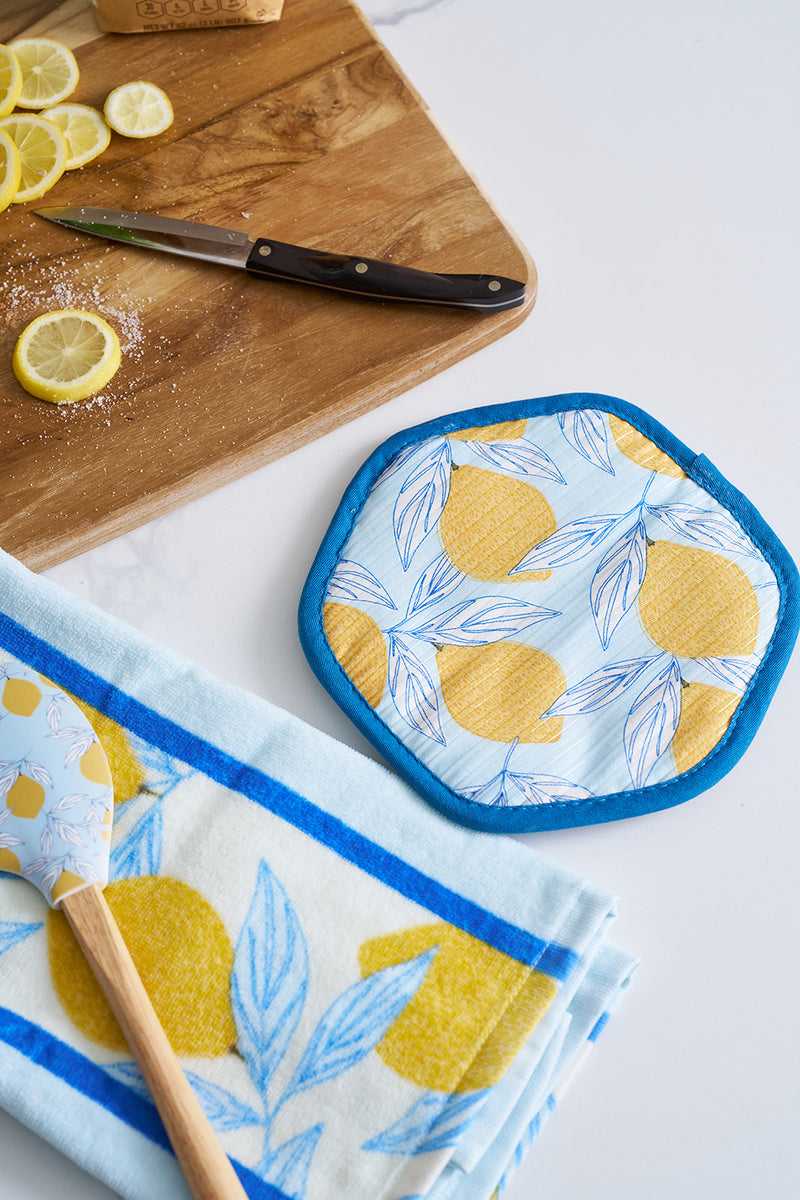 A kitchen scene showcases a cutting board with sliced lemons and a knife beside the Lots of Lemons Pot Holder. A lemon-patterned towel complements the decor, accompanied by a wooden spoon featuring a lemon design on its handle.