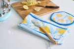 A kitchen counter showcases a cutting board with lemon slices and a knife. Next to it, the Lots of Lemons Pot Holder with a silicone grip matches the blue and yellow lemon-patterned towel and spatula, while part of a metal appliance is visible nearby.